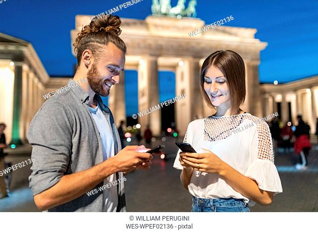 Happy couple using smartphones at Brandenburg gate at blue hour, Berlin, Germany