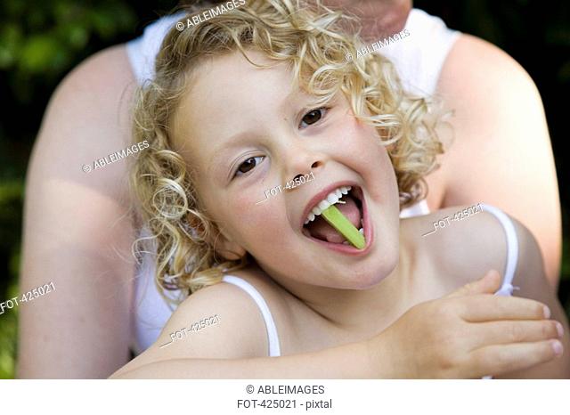 Young girl sitting on her mother's lap with a piece of celery in her mouth