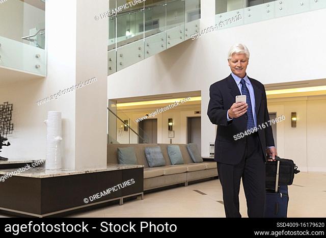 Mature Caucasian man looking at cell phone while walking through hotel lobby with luggage
