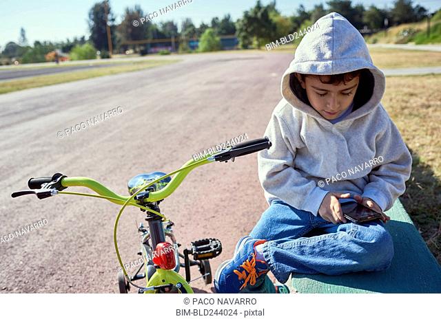 Hispanic boy sitting near bicycle texting on cell phone
