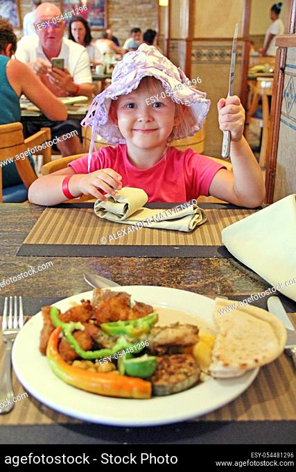 Little girl eating in restaurant. Baby with knife in hand. Happy girl emotionally waiting for dish in restaurant. Little girl having lunch in restaurant with...