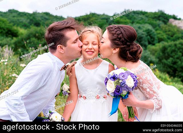 young parents on a wedding walk in wedding dresses kiss their young daughter in cheeks. Concept of a happy young family