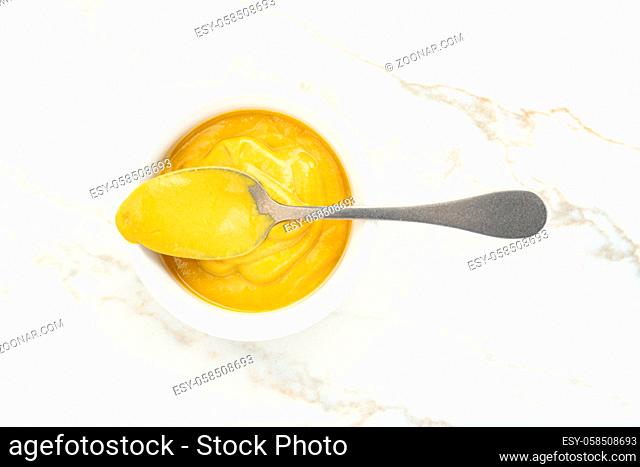 Mustard sauce in a bowl and spoon on white marble table. Top view