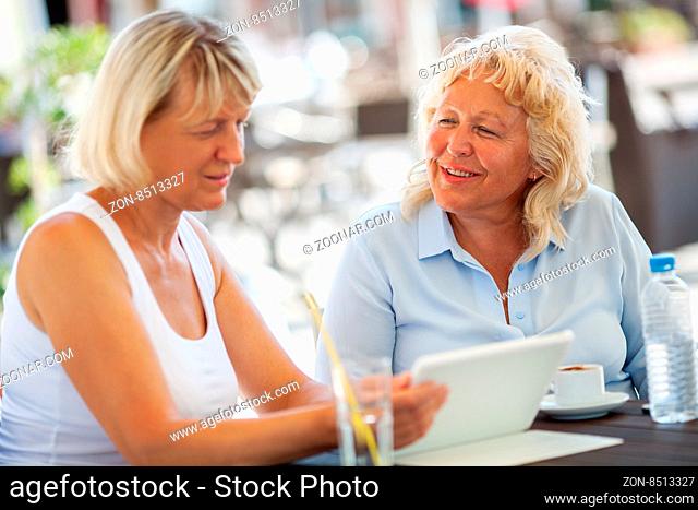 Happy two women of senior age having drinks and working with tablet PC in outdoor cafe on bright summer day
