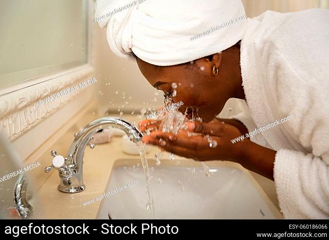 African american woman in bathroom with towel on head, standing at basin washing face