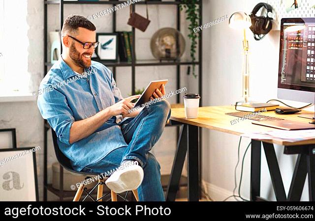 Side view of cheerful young male in casual wear and glasses using tablet while sitting with takeaway coffee at workplace with large monitor