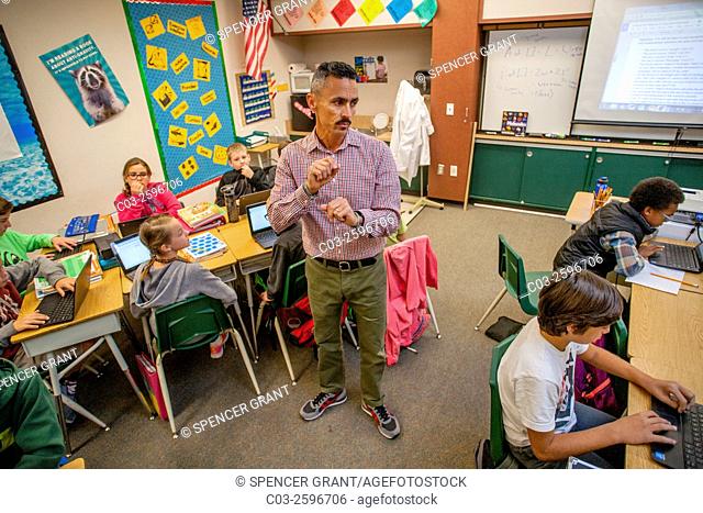 A Hispanic 5th grade elementary school teacher in San Clemente, CA, watches his multiracial students using Google Chromebook laptop computers in the classroom