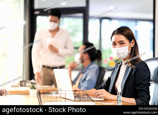 Portrait of asian office employee businesswoman wear protective face mask work in new normal office with interracial colleague in background as social distance...