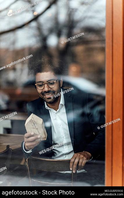 Smiling male entrepreneur looking at house model while sitting in cafe seen through glass