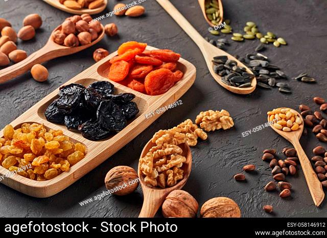 Various varieties of nuts, seeds and dried fruits lying in wooden spoones and dish on black slate background. Top view. Healthy food