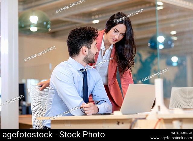 Female professional looking at laptop by male colleague at desk in coworking office