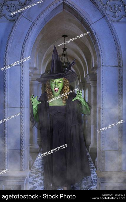 Young girl dressed as witch making scary face to camera standing in front of smokey stone hallway