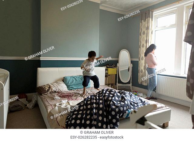 Little boy is jumping on his mother's bed, who is standing by the window with her baby son in her arms