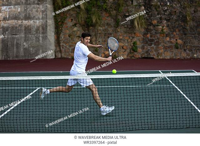 Side view of a young Caucasian man playing tennis, running up to a ball with a wall behind him
