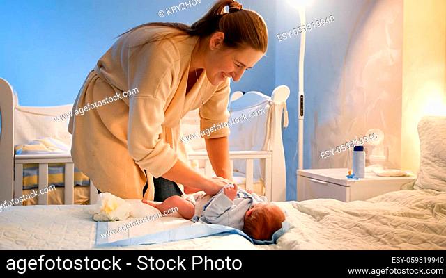 Smiling young mother changing diapers and wiping her newborn baby son got messy on bed at night. Sleepless nights of parents and baby hygiene