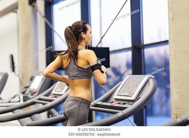 sport, fitness, lifestyle, technology and people concept - woman with smartphone or player and earphones exercising on treadmill in gym