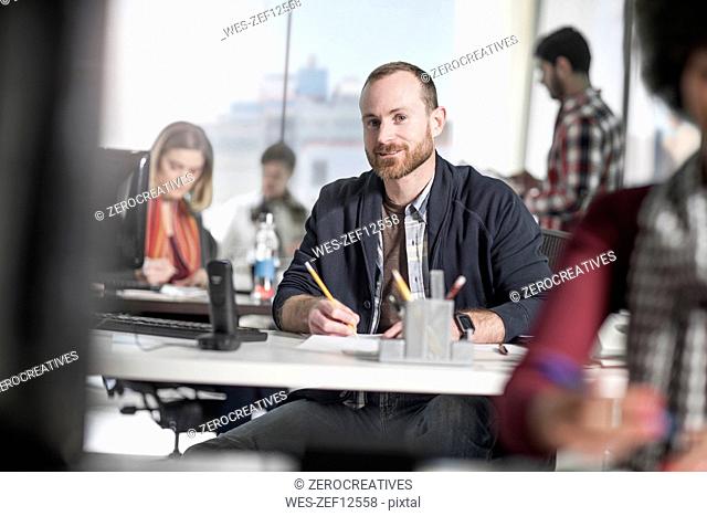 Portrait of smiling man working at desk in office surrounded by coworkers