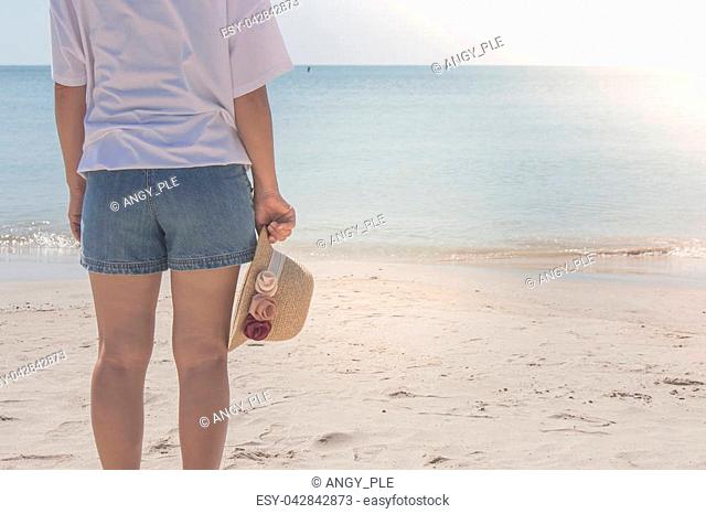 Woman wearing white t-shirt, she standing on sand beach and holding weave hat in hand, she looking at the sea and blue sky