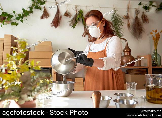 Woman with protective eyewear and mask mixing ingredient in bowls while making soap in workshop