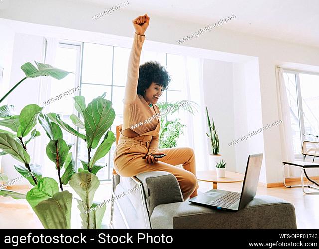 Excited woman sitting with hand raised looking at laptop on sofa