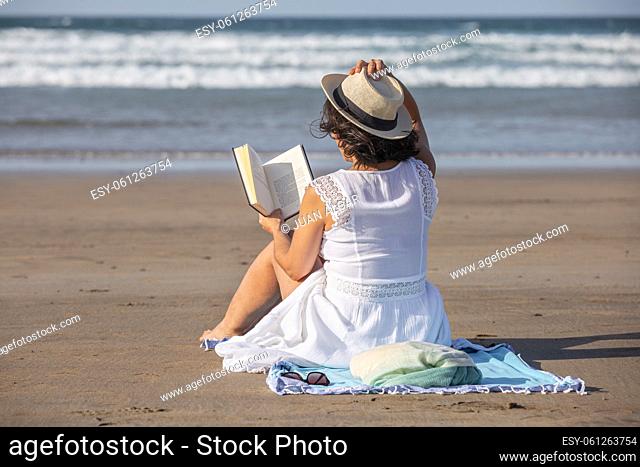 Back view of female traveler in white dress touching hat and reading interesting book while sitting on sandy beach near waving sea during summer vacation