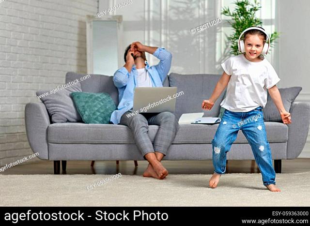 Cheerful playful kid dancing and unhappy busy father working with laptop at home