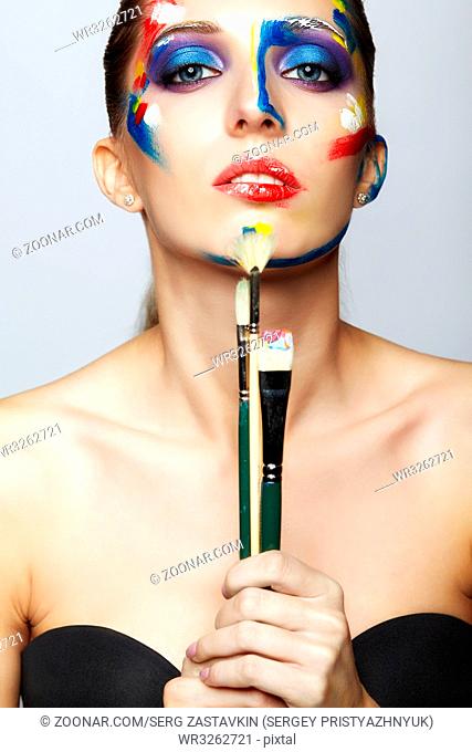 Young woman painter with paint brush in hand and acrylic paint on face