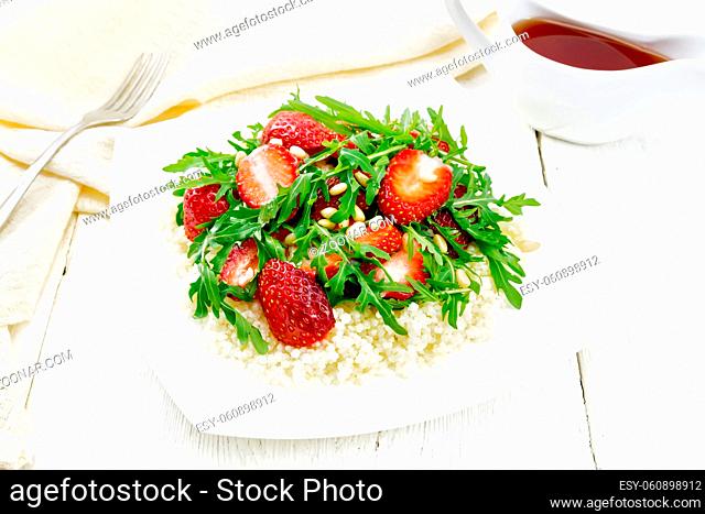 Strawberry, couscous, cedar nuts and arugula salad dressed with balsamic vinegar and olive oil in plate, a towel and a fork on the background of light wooden...