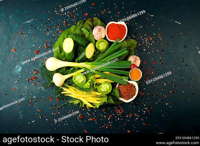 FRESH VEGETABLES AND SPICES ON A ROUND WOODEN BOARD ON A DARK STONE BACKGROUND. THE CONCEPT OF VINTAGE. SPICES THE YOUNG CORN ASPARAGUS GREEN TOMATO CUCUMBER ON...