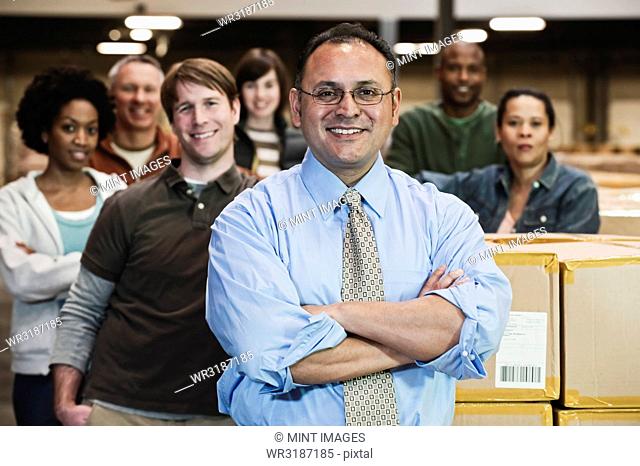 Team portrait of multi-ethnic warehouse workers lead by a Hispanic American male executive and surrounded by large racks of products stored in cardboard boxes...