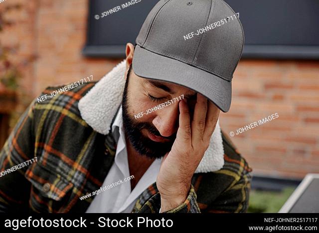 Man with beard and baseball cap touching face