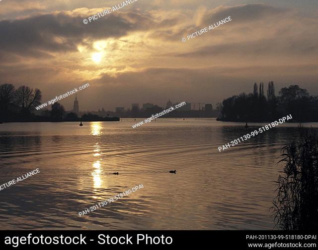 27 November 2020, Brandenburg, Potsdam: The setting sun can be seen in a gap in the clouds and reflects its light in the water of the deep lake through which...