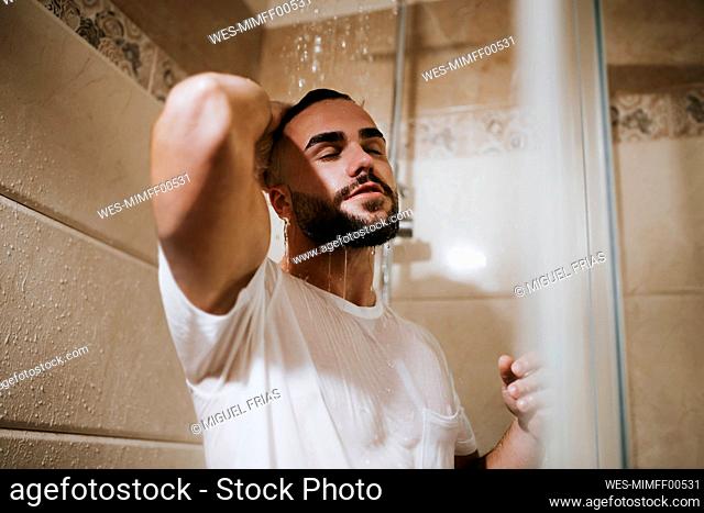 Young man in t-shirt with hand in hair taking shower in bathroom at home