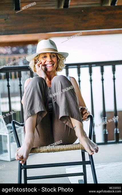Portrait of a mature caucasian blonde young woman in her 50s talking on a mobile phone smiling and wearing a hat and sitting in a relaxed pose in a rocking...