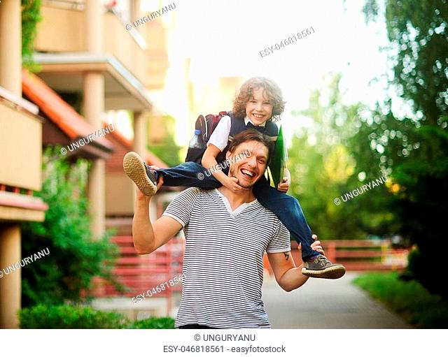 The father carries on his shoulders of his son-schooboy. Little learner with one arm hugs dad for the neck and the other holding a green folder