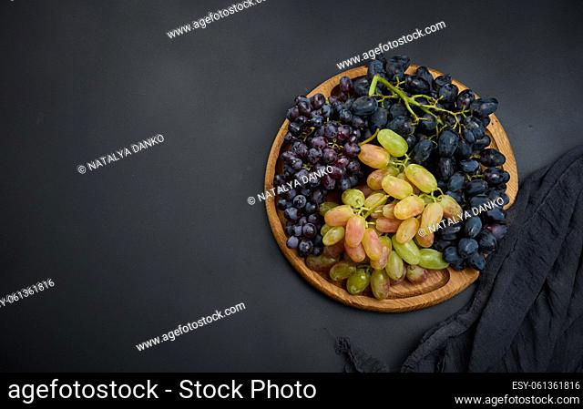Black and green grapes in a wooden round plate on a black table, top view