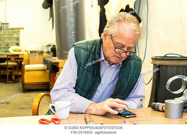 Tilburg, Netherlands. The 72 year old upholsterer Boy, reading the messages on his smartphone, while sitting at his workbench