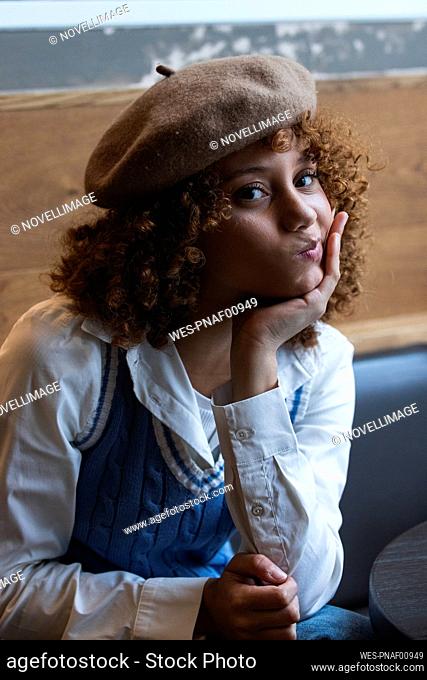 Teenage girl wearing beret making face while sitting with hand on chin at cafe