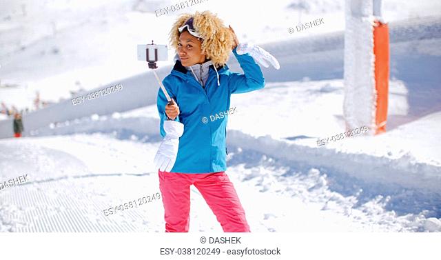 Smiling young woman posing for a photograph waving at her mobile phone camera as she takes a selfie on a stick in thick white snow