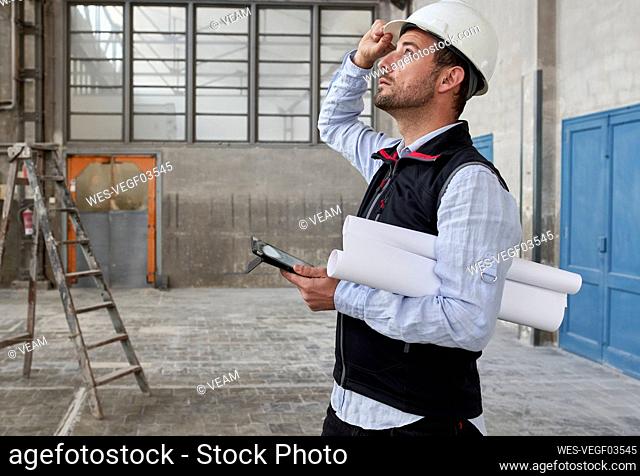 Male architect with digital tablet and blueprints looking up while standing in building