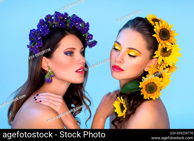 Two beautiful young women with yellow and violet flowers in hair. Bright makeup. Beauty shot. Copy space. Over blue background