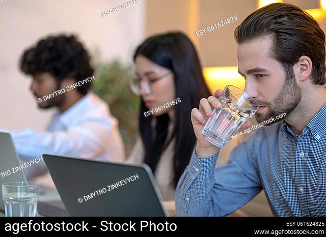 Office employee seated beside his busy coworkers staring at his laptop and drinking a glass of water