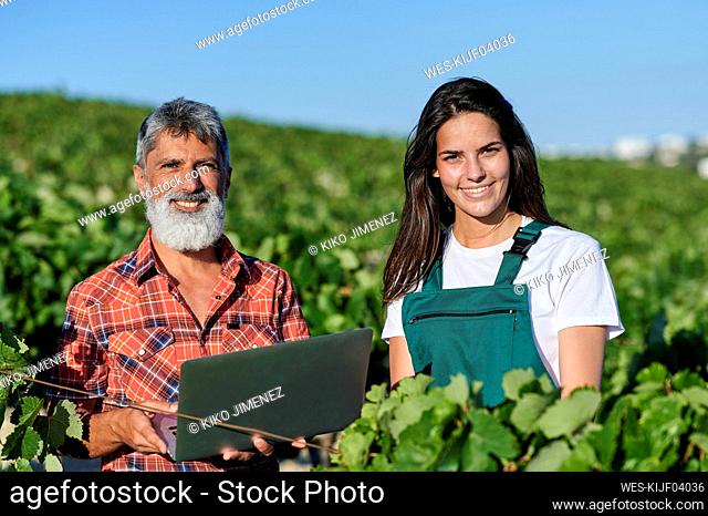 Smiling male and female farmers standing amidst plants in vineyard on sunny day
