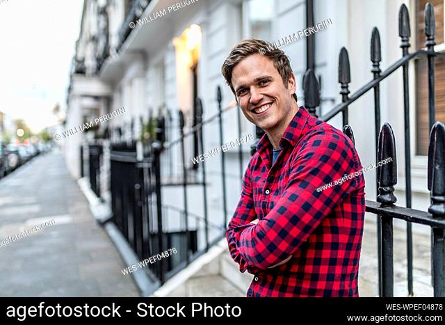 Smiling man with arms crossed leaning on metal fence at footpath