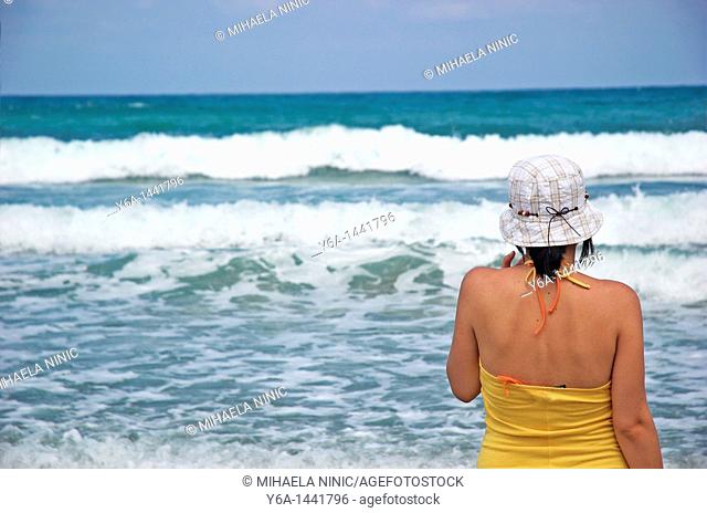 Woman looking out towards sea on beach wearing hat with waves crashing
