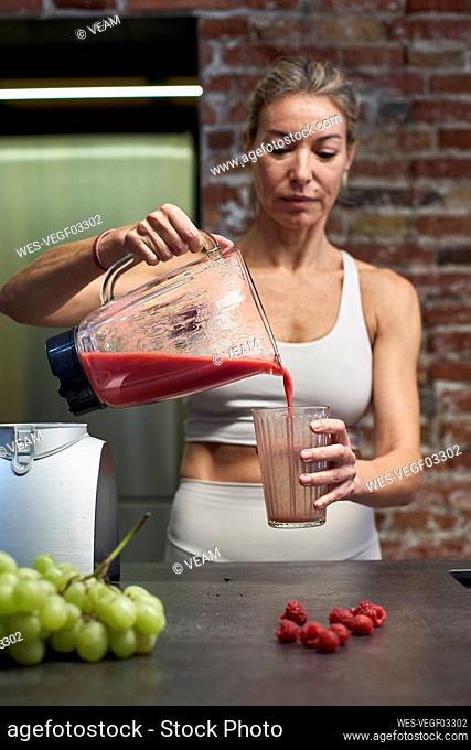 Mature woman pouring raspberry fruit smoothie in glass at kitchen
