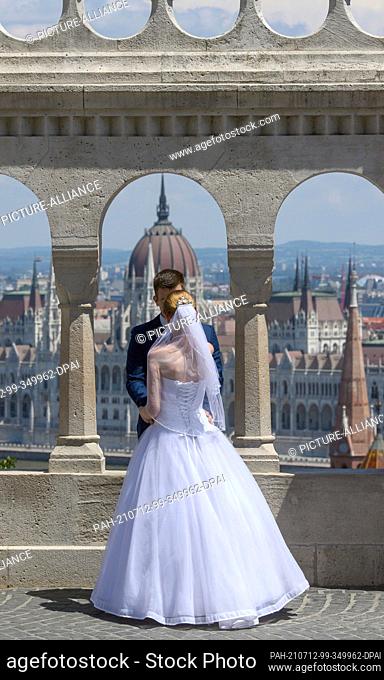 26 June 2021, Hungary, Budapest: A wedding couple stands by the arches of the Fishermen's Bastion with the Parliament building on the Danube in the background