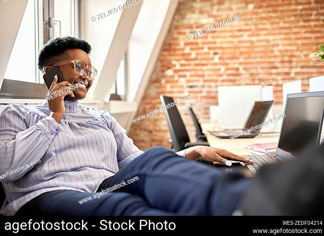 Female professional wearing eyeglasses talking on smart phone while using laptop at office
