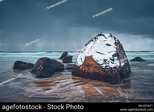 Rocks covered with snow on Norwegian sea beach in fjord in stormy weather with clouds. Skagsanden beach, Flakstad, Lofoten islands, Norway