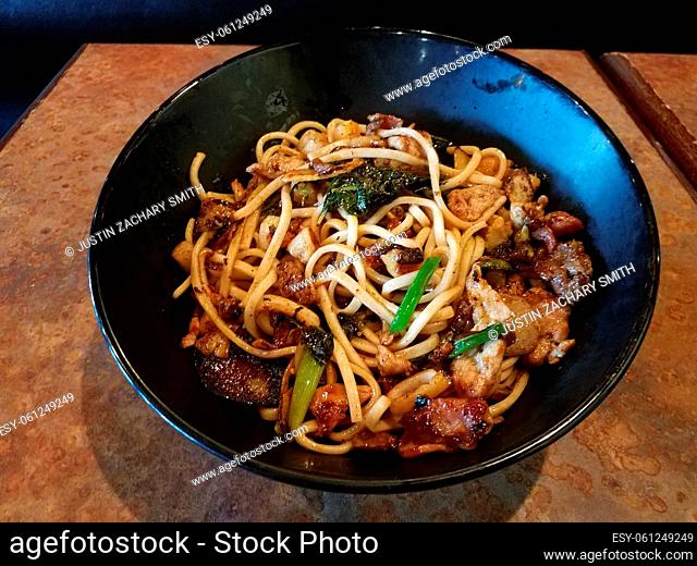 asian noodles in black bowl with beef, pork, bacon, and vegetables on table